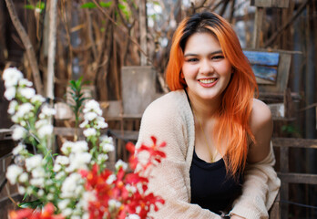 Asian happy beautiful cheerful outdoor portrait woman light orange hair smiling warmly among blooming flowers, Natural wooden textures greenery create cozy rustic garden joyful fresh nature concept.