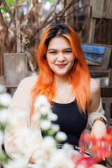 Asian happy beautiful cheerful outdoor portrait woman light orange hair smiling warmly among blooming flowers, Natural wooden textures greenery create cozy rustic garden joyful fresh nature concept.