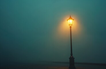 Old retro street lamp stands tall, shines bright yellow light on dark foggy night. Vintage lantern illuminates empty urban street path, creating warm ambiance. Solitary light post against deep blue