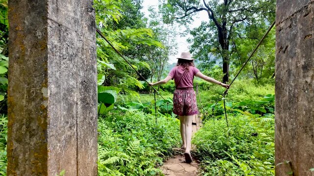 Woman in a hat walking on a simple suspension bridge in a lush tropical jungle. slow motion