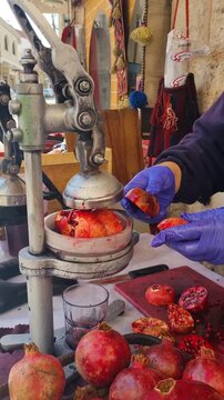 Vertical close-up of a vendor wearing gloves extracting fresh pomegranate juice using a hand-operated press outside his shop in Madaba&rsquo;s old town on a sunny day with lively street activity, Jordan