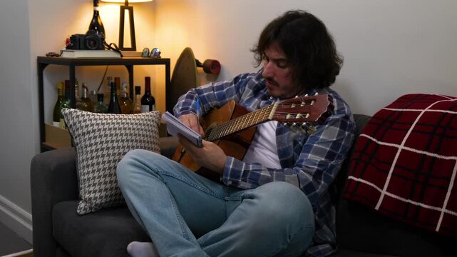 A man sits on the sofa writing music using a guitar, notebook and pen.