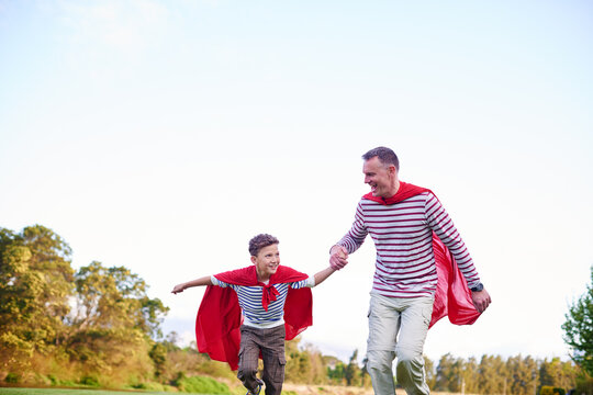 Park, child and dad with superhero costumes, family and fun with imagination, bonding and happiness. Outdoor, man and son with cape, pretend and holding hands with boy in nature, mockup space or play - Powered by Adobe