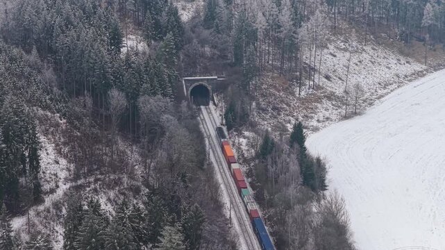 A freight train enters a tunnel in a winter landscape. A row of colorful containers traveling along the tracks.