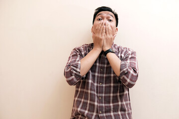Southeast Asian man wearing flannel shirt showing shocked face expression, with hand covering mouth, on white isolated background.