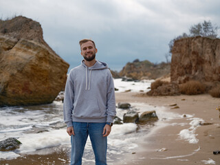 Smiling Man Standing on Wild Sea Coast in Hooded Sweatshirt