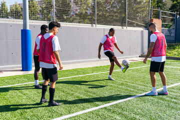 Four young men training on an artificial turf field, one player juggling a football with his foot, others watching