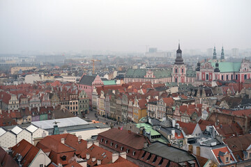 Obraz premium Wide panoramic view of Poznań Old Town with colorful historic buildings, red rooftops and church towers on a misty day.