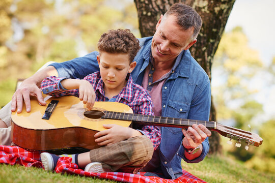 Playing, guitar and father with kid in nature for learning, bonding or musical hobby together. Happy, love and dad teaching boy child song with acoustic string instrument on picnic outdoor in park. - Powered by Adobe