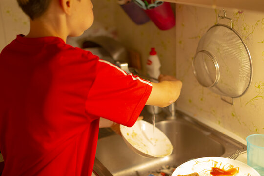 teen washing dishes at sink in red shirt, scrubbing plate under running faucet with soap suds and sponge, countertop cluttered with bowl and cup, warm afternoon light casting soft shadows, candid