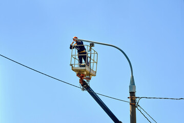Worker in helmet and safety harness uses crane lift to install led street lamp during outdoor...