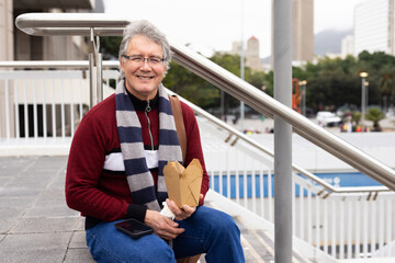 Man in maroon sweater sitting against steel railing in plaza holding takeout box and smartphone