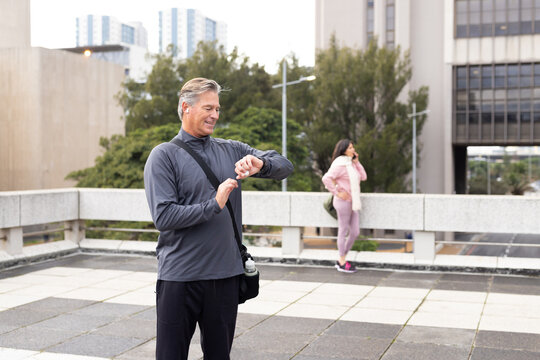 Senior couple exercising on urban terrace, checking smartwatch and talking on smartphone
