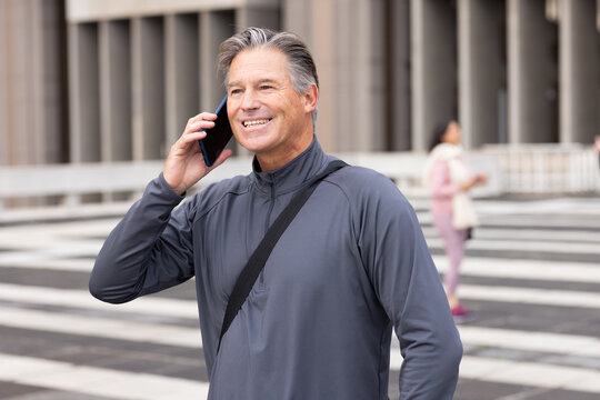 Man wearing gray jacket with crossbody strap talking on smartphone on plaza in front of columns