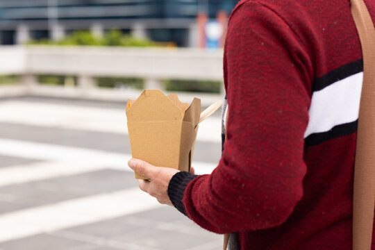 Mature adult male standing at pedestrian crossing holding takeout box with chopsticks, copy space