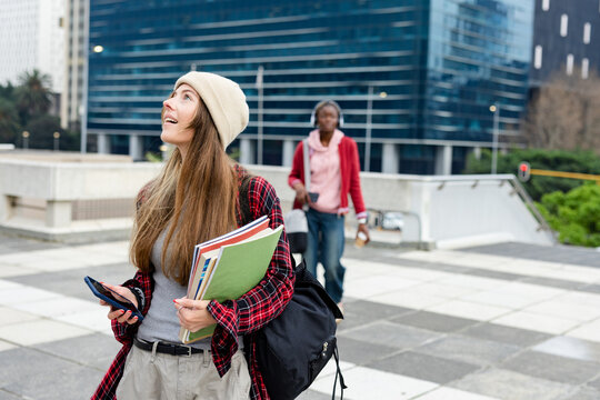 Diverse female students standing on plaza by glass office building using smartphone, copy space