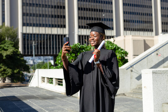 African American woman wearing gown cap posing on campus taking selfie with diploma and smartphone