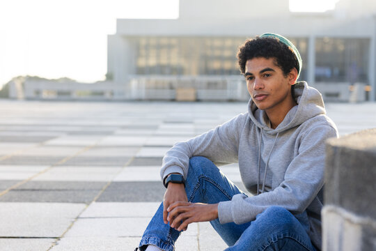 Hispanic male student sitting on stone ledge at urban plaza tiles, checking smartwatch, copy space