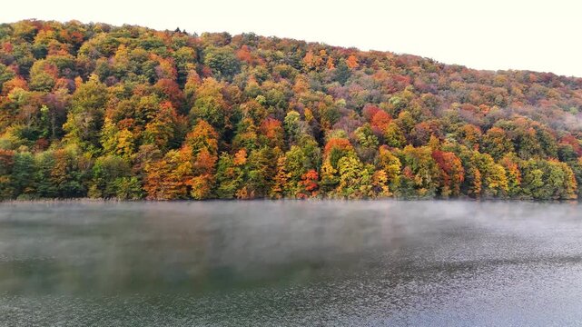 Mist drifts above Lac du Val as autumn colors blanket the forested hillside near Le Herisson in France. Vibrant foliage in orange, red, and green surrounds the calm lake, creating a seasonal landscape