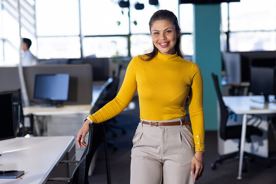 Woman wearing mustard-yellow sweater, beige-trousers leaning on office chair in open-plan office