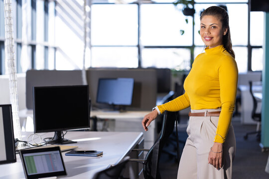 Businesswoman wearing yellow top standing in office beside dual monitors and laptop, copy space