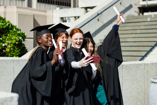 Diverse female graduates posing on campus in caps holding diplomas taking selfie with smartphone