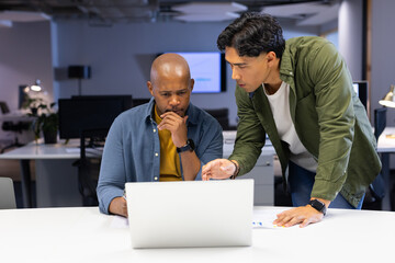 Diverse male coworkers collaborating over laptop at office wearing mustard T-shirt and olive jacket