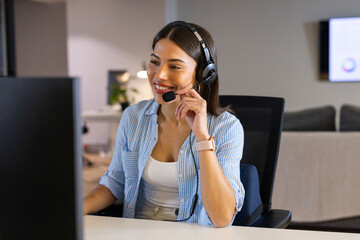 Indian woman office worker speaking through headset at open-plan office desk with computer monitor