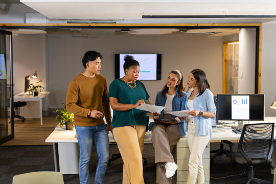 Diverse coworkers discussing project details at open-plan office with display screens and reports