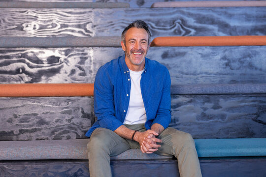 Mature man sitting and smiling on wooden bench at lecture hall wearing blue shirt, beaded bracelet