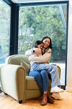 Diverse mother and daughter hugging on green armchair in living room, holding smartphone on armrest