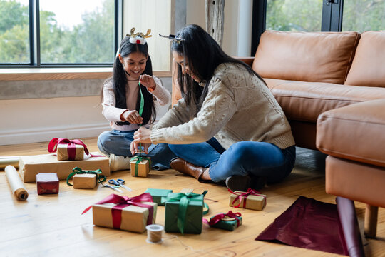 Diverse mother and daughter sitting in living room wrapping gift boxes with kraft paper and ribbons