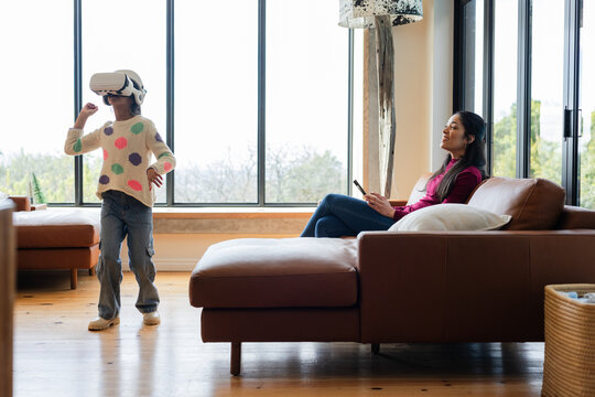 Diverse mother and daughter playing virtual reality game in living room using VR headset and tablet - Powered by Adobe