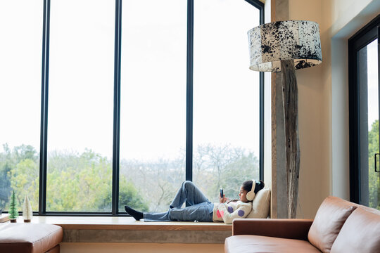 Indian girl reclining on window seat in home lounge using smartphone with headphones, copy space