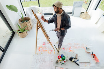 Indian man painting on canvas at easel in bright home studio with paint jars and palette