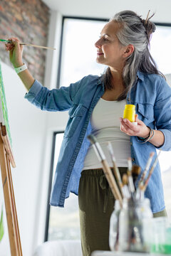 Painter holding paintbrush and yellow paint jar applying green paint to canvas on easel in studio