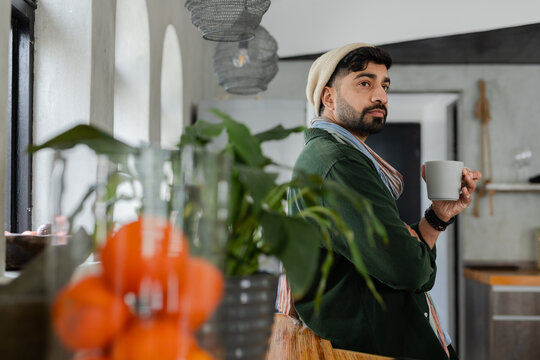 Indian man leaning at wooden counter in kitchen holding white mug wearing knit beanie, copy space