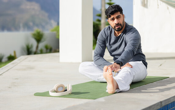 Indian male in sportswear stretching on yoga mat at terrace by lake with headphones, copy space