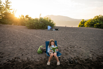 A young girl reads a book on a sandy beach at sunset