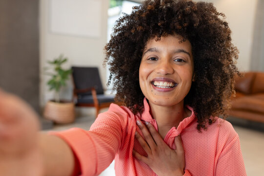 African American woman sitting at home extending arm taking selfie with smartphone in coral top