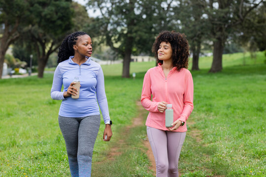 Diverse female friends in athletic wear walking on path in park with water bottles and smartwatches