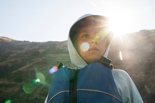 A young boy in a life jacket looks into distance backlit by sun