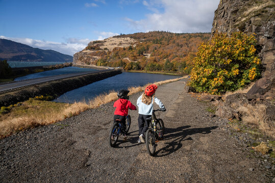 A young boy and young girl walk bikes on a trail by wide river.