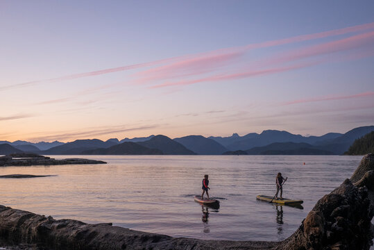 A young girl and young boy paddle boards at dusk among mountains