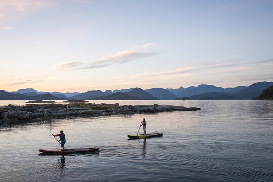 A young girl and young boy paddle boards at dusk among mountains