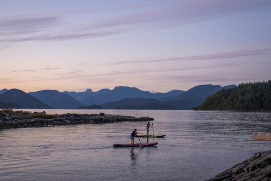 A young girl and young boy paddle boards at dusk among mountains