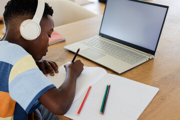 African American boy in striped shirt writing in notebook at home desk with laptop, pencil, headset
