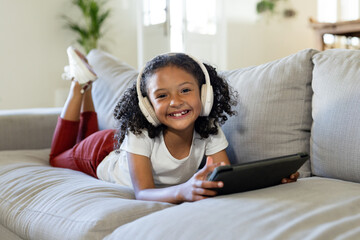 Female child lying on light grey couch in living room holding black tablet wearing headphones