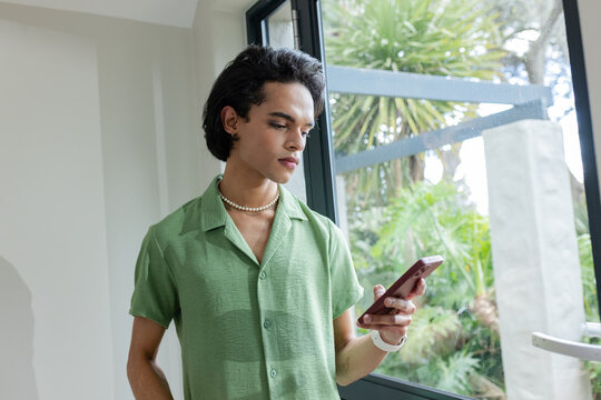 Male wearing pearl necklace and smartwatch checking smartphone at window in modern home, copy space