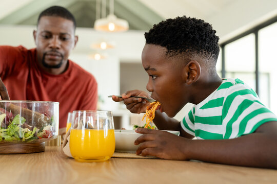 African American father and son serving salad while son eating spaghetti at dining table at home - Powered by Adobe